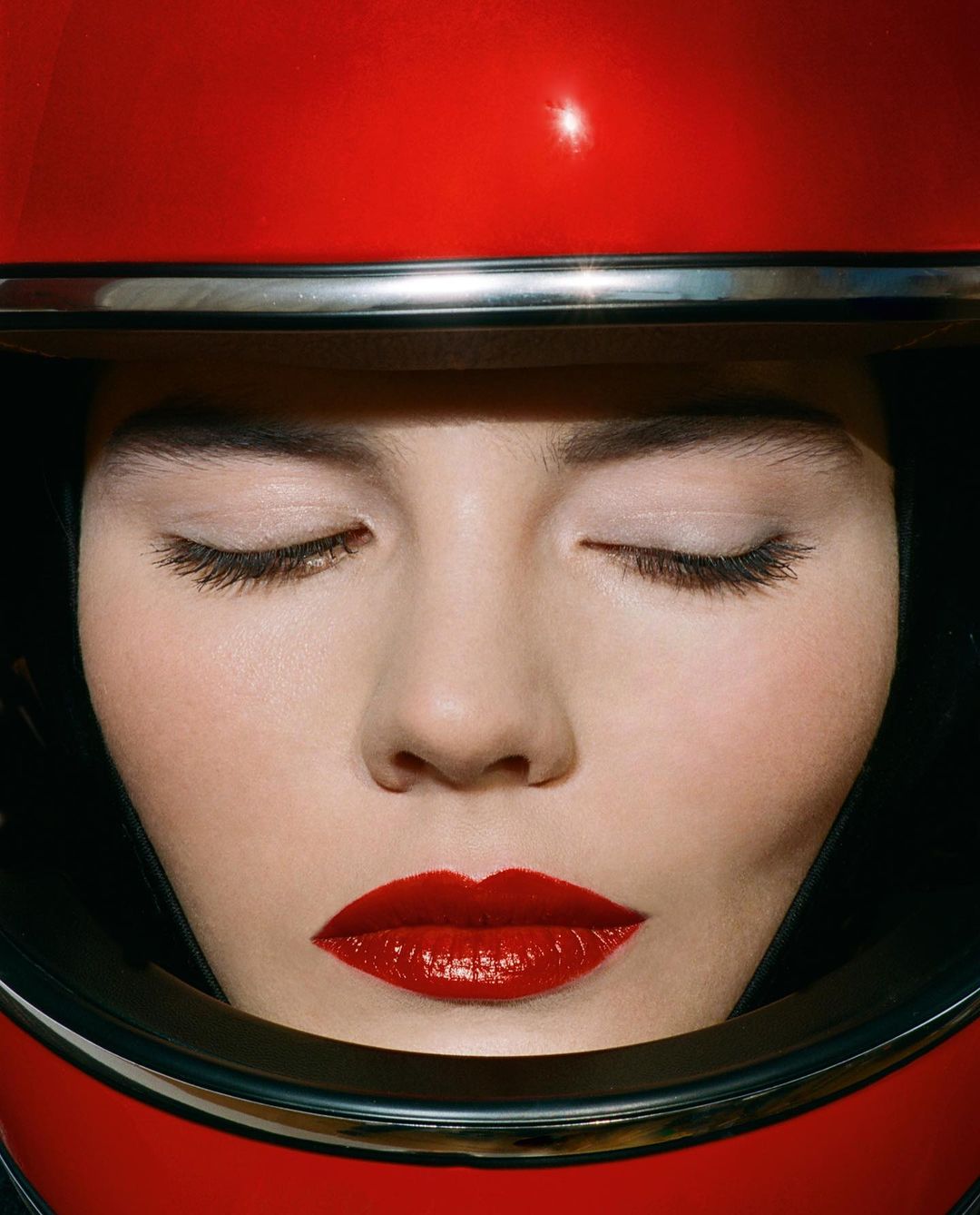 Woman with closed eyes, wearing red helmet and red lipstick in a close-up studio shot, focusing on the makeup and helmet's reflective surface.