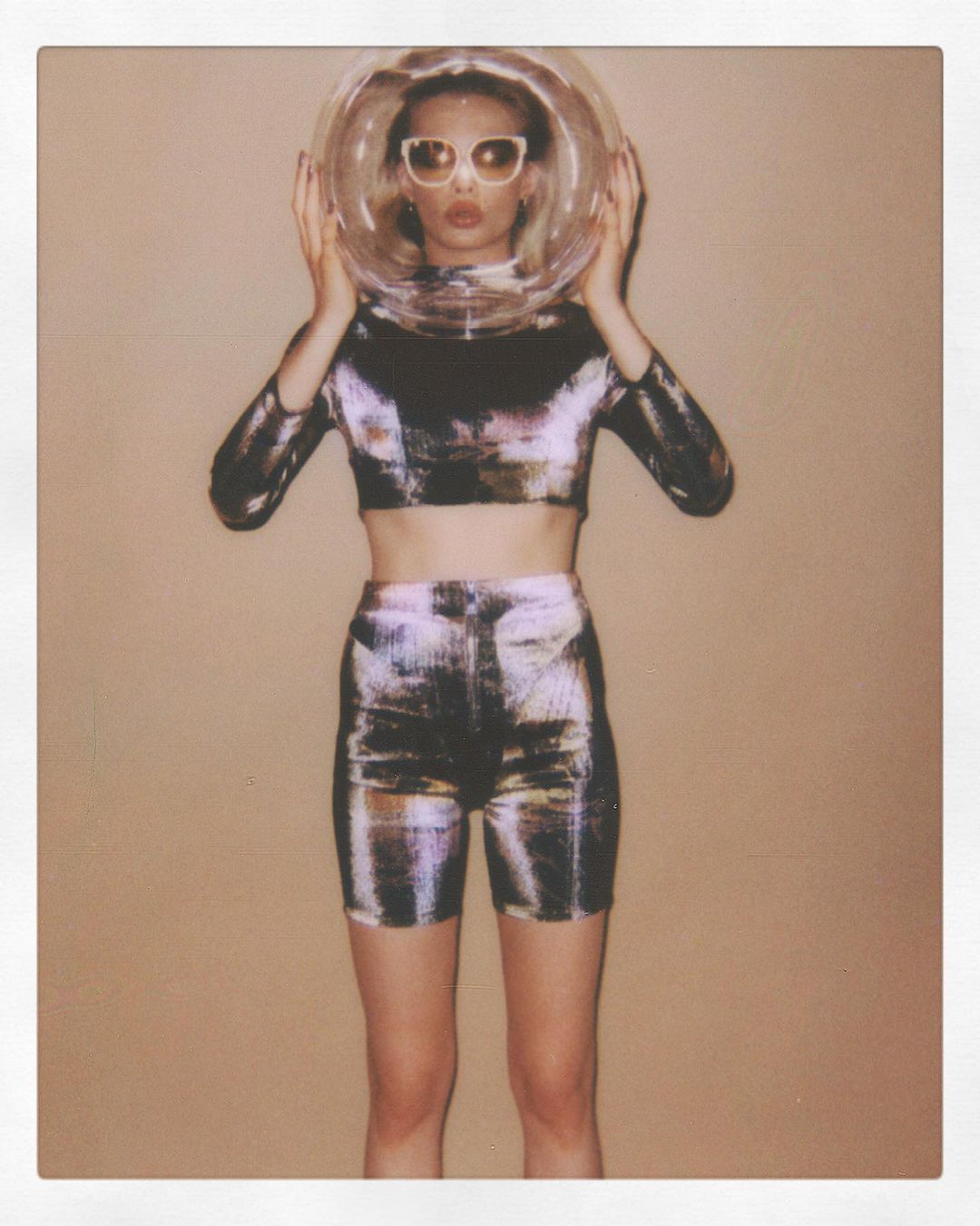 Young woman in futuristic fashion wearing sunglasses and a clear space helmet against a plain backdrop in a studio setting.
