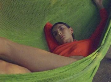 Eye-level portrait of a young adult woman resting in a green mesh hammock, wearing a red top, looking slightly upward.