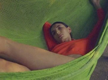 Eye-level portrait of a young adult woman resting in a green mesh hammock, wearing a red top, looking slightly upward.