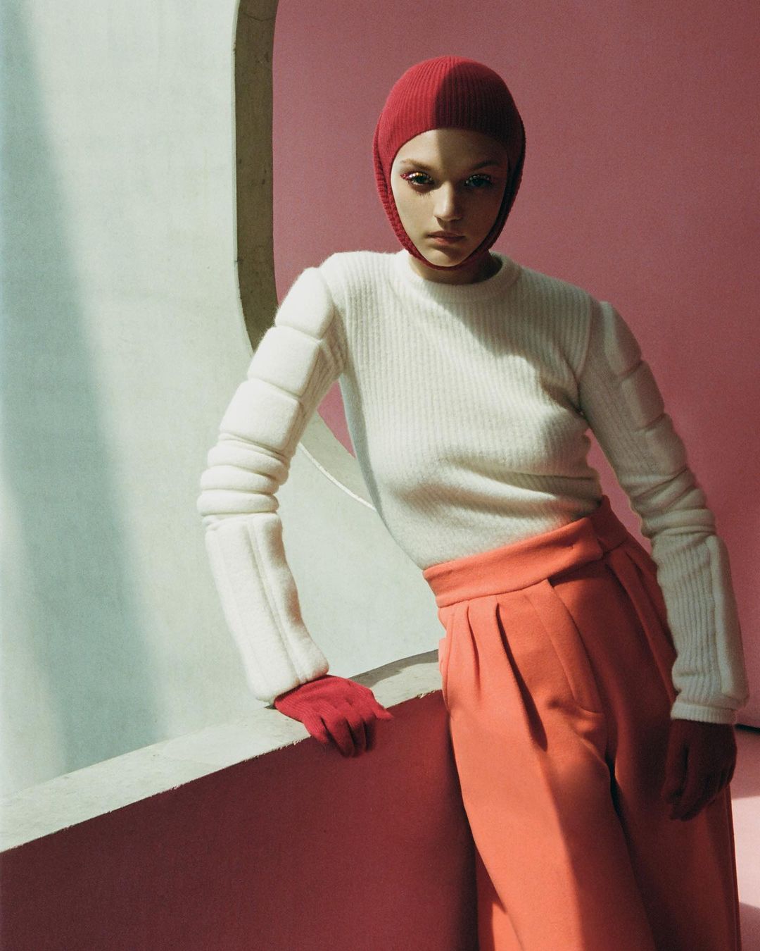 Fashion shot of a woman wearing a white sweater, orange pants, and a red head covering, posing in front of a pink wall with architectural elements.