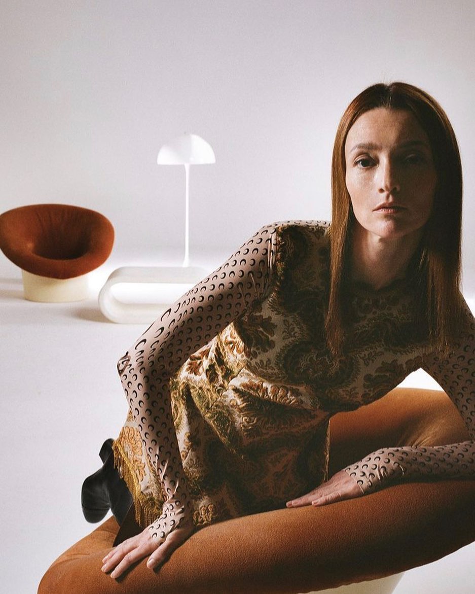 A model poses indoors wearing a patterned dress with matching long sleeves, sitting on a brown chair with a white table and lamp behind.