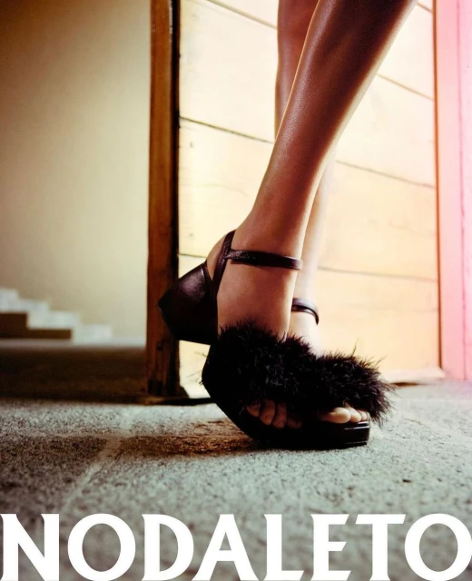 Woman's legs and feet wearing black strappy platform sandals with fur detail, standing near a doorway with stairs in the background.