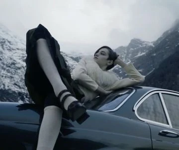 Fashion shot of a woman in vintage style clothing sitting on a classic car with a mountain backdrop, overcast day.