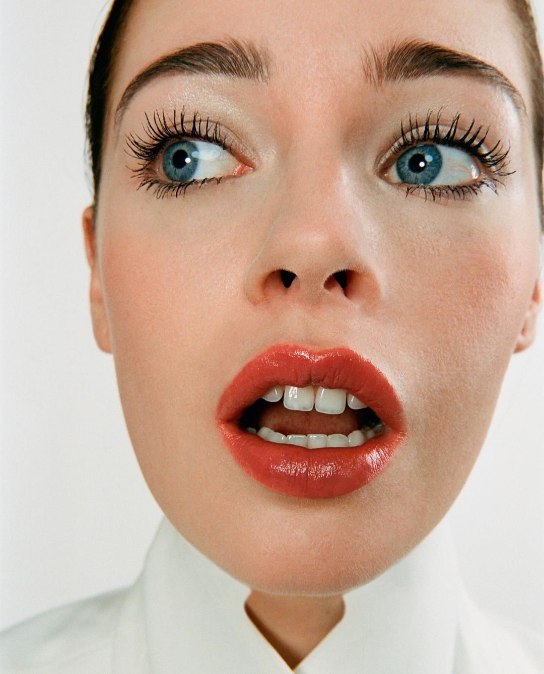 Close-up portrait of a young adult woman with blue eyes and coral lipstick, looking off-camera with an open mouth against a white background.