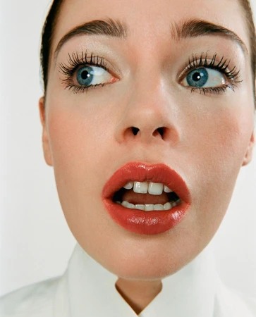 Close-up portrait of a young adult woman with blue eyes and coral lipstick, looking off-camera with an open mouth against a white background.