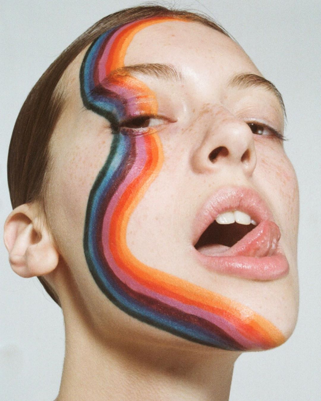 Woman with colorful rainbow face paint, mouth open and tongue sticking out against a light gray backdrop in a beauty shot.