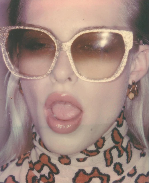 Close-up of a young woman with blonde hair wearing sunglasses, earrings, and a patterned top, sticking her tongue out at the camera.