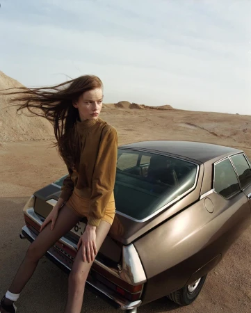 Model with long blowing hair sitting on the back of a brown car in a desert landscape by Damien Krisl