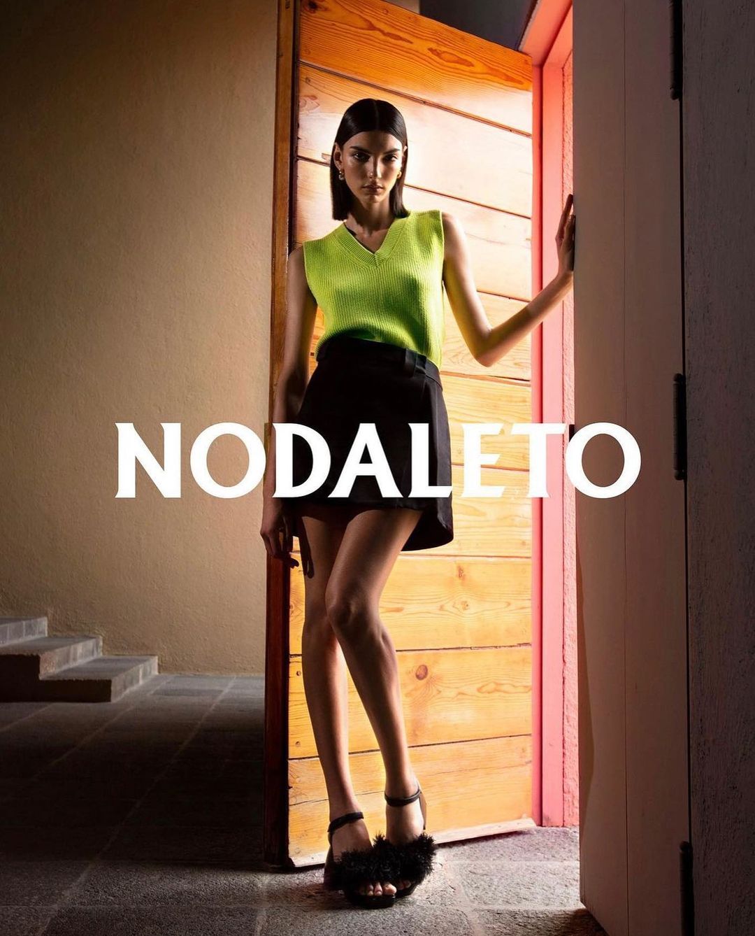 An adult woman poses near a wooden door, wearing a neon green top, black skirt, and black furry heels, set against a tan wall.