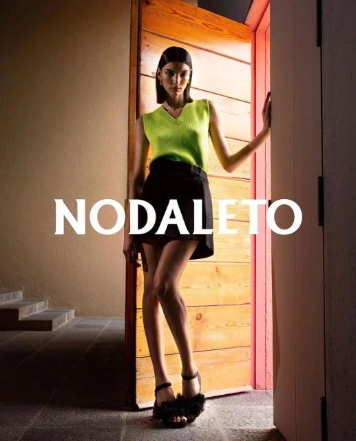 An adult woman poses near a wooden door, wearing a neon green top, black skirt, and black furry heels, set against a tan wall.