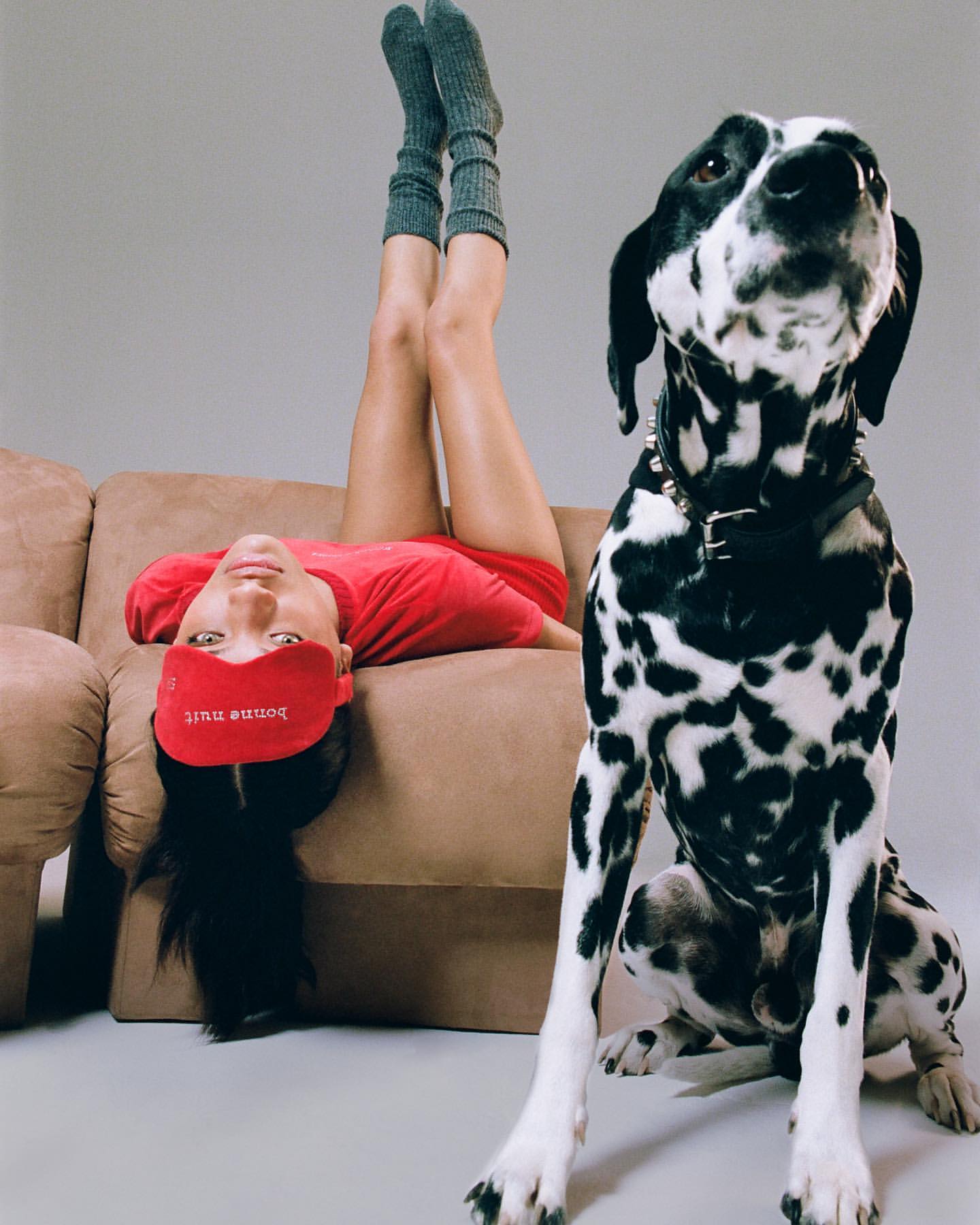 Woman lying on a couch with legs up, wearing a sleep mask, next to a Dalmatian dog with a studded collar against a grey background.