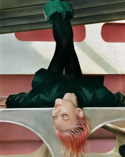 Upside-down shot of a woman with pink hair in a green jacket and black leggings, legs are raised, resting on a horizontal plane.