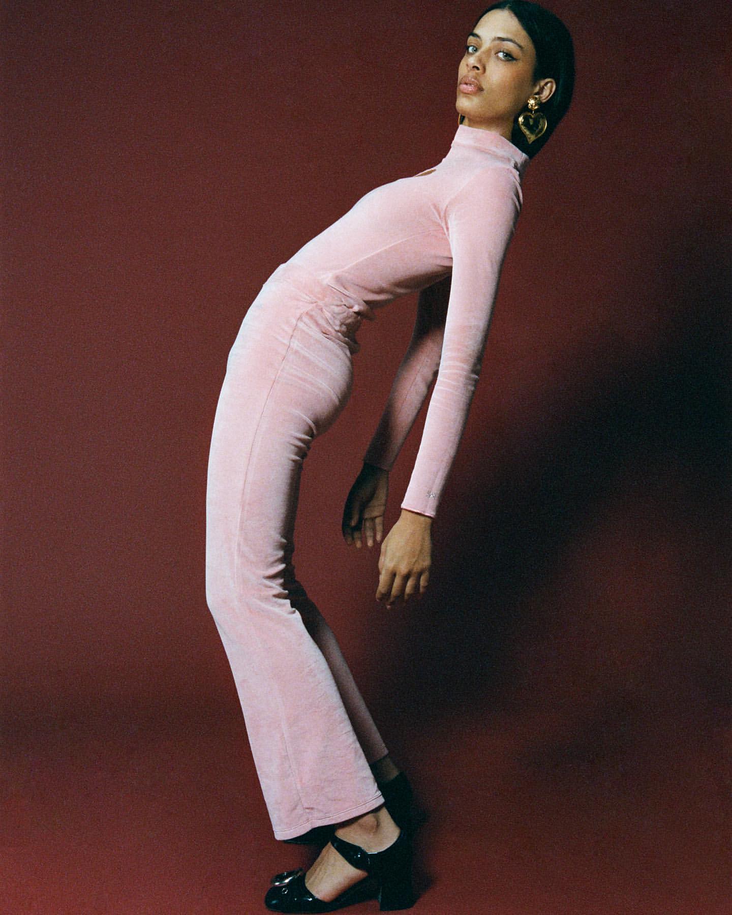 Woman in pink velvet turtleneck dress and black heeled shoes in studio setting. Fashion editorial. Portrait shot on red background.
