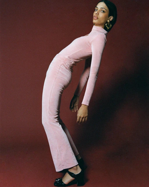 Woman in pink velvet turtleneck dress and black heeled shoes in studio setting. Fashion editorial. Portrait shot on red background.
