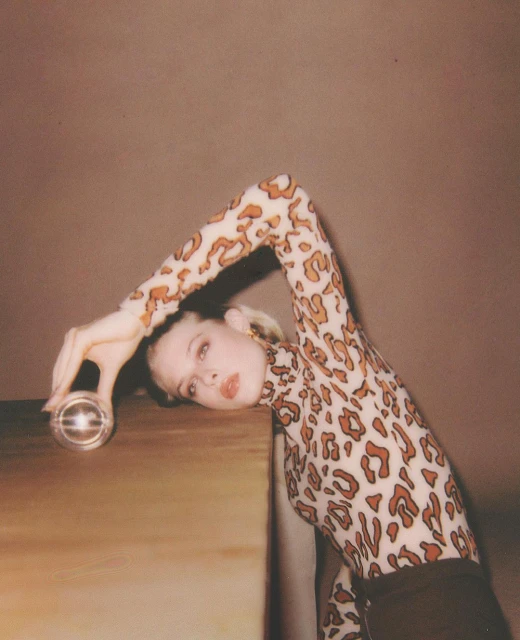 Woman in leopard print top leans on table with glass orb. Fashion portrait with soft lighting and warm tones.