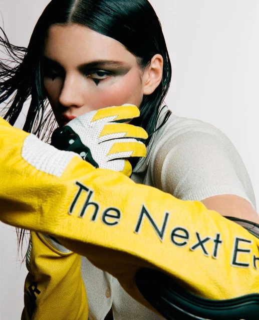Editorial shot of a woman with black hair and makeup, wearing white top and yellow racing jacket and gloves in a brightly lit studio.