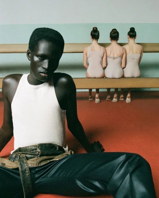 A dark-skinned woman in a white tank top and dark pants sits on a red floor, with three ballerinas on a bench in the background.