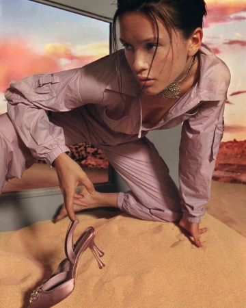 Model in pink jumpsuit kneeling on sand with matching heeled sandal against sunset backdrop