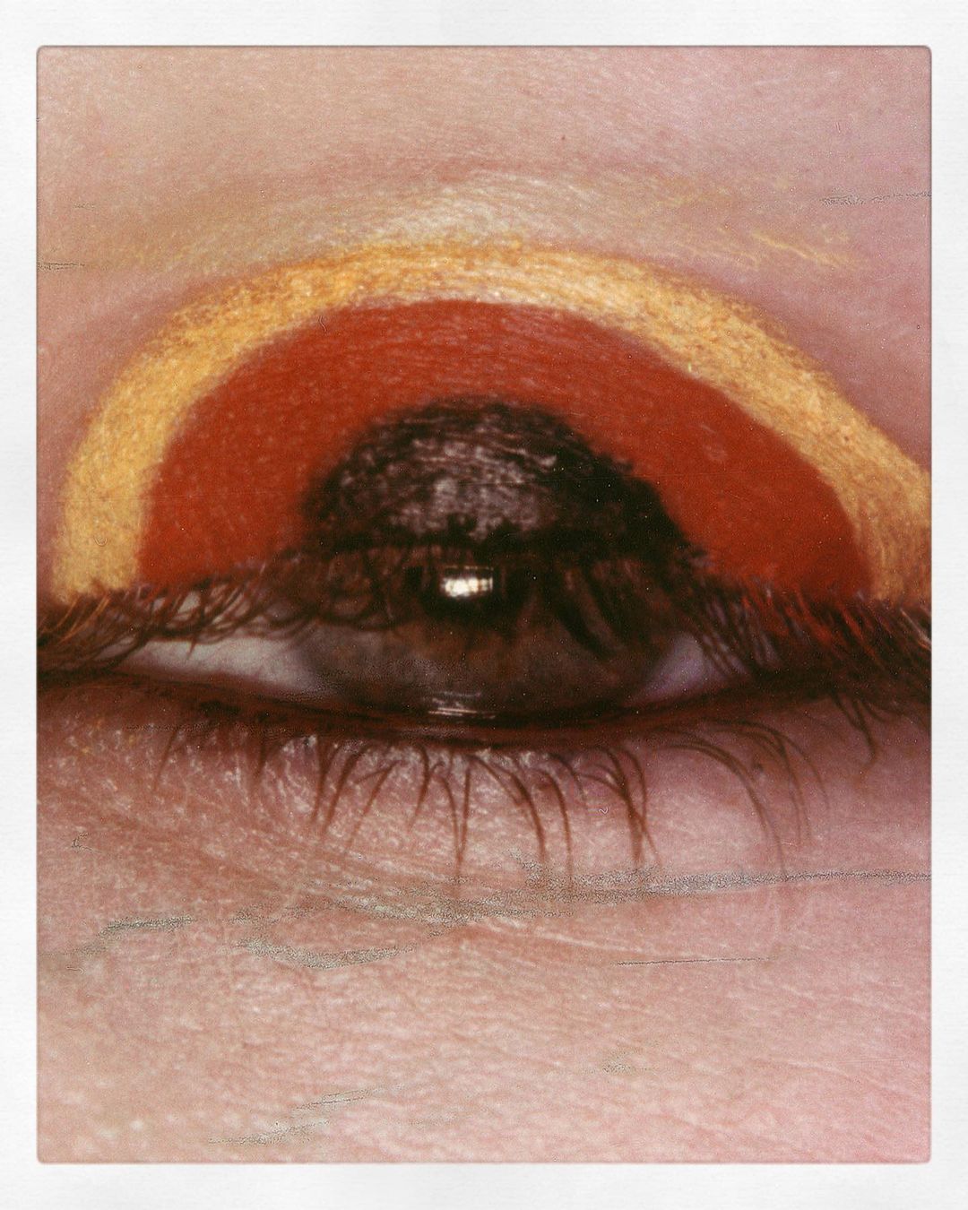 Close-up of a woman's eye with colorful eyeshadow makeup in hues of gold, red, and brown creating an artistic arc above the eye.