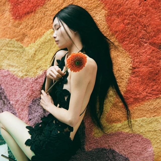 Woman with long black hair reclines on a vibrant, multicolored shag carpet, holding an orange flower. She is wearing a black floral dress.