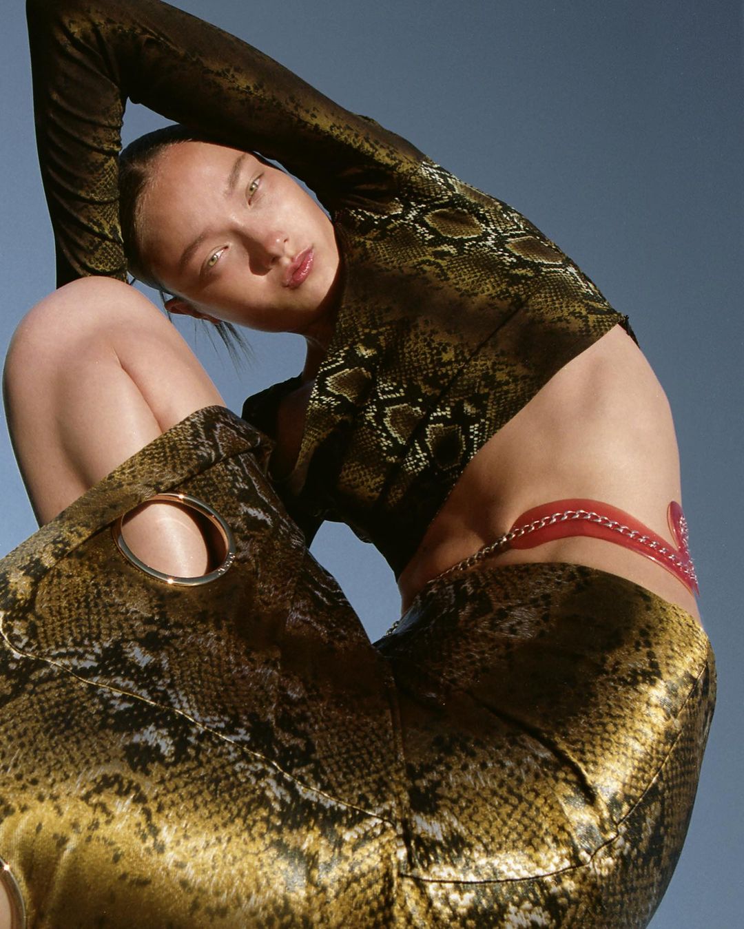 A young woman in a snakeskin print top and skirt poses against a blue sky in a fashion-forward style. She has a confident expression.