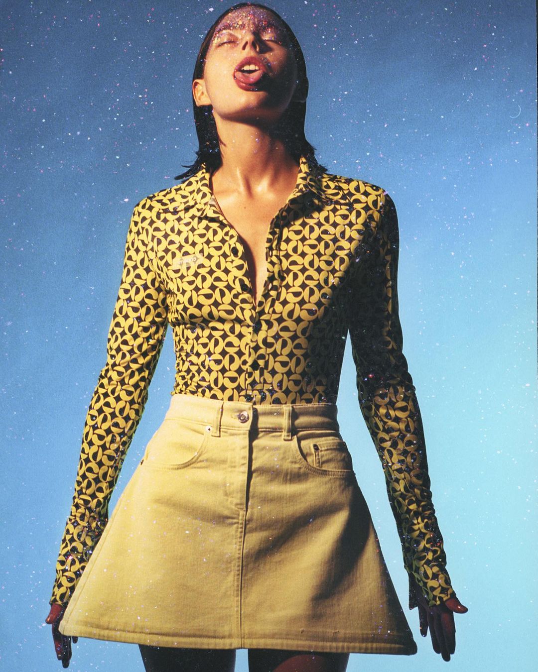 Woman in patterned top and yellow skirt poses against a speckled blue backdrop with glitter on her face and her tongue slightly out.