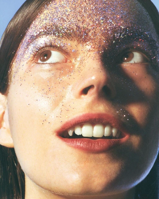 Glamorous close-up shot of a woman's face with sparkling pink glitter makeup, upward gaze, illuminated by soft, natural light.