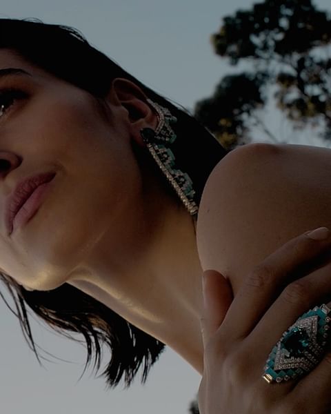 Close-up of a dark-haired woman with large gemstone earrings and a ring with a large gemstone, outdoors against a soft blue sky.