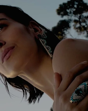 Close-up of a dark-haired woman with large gemstone earrings and a ring with a large gemstone, outdoors against a soft blue sky.