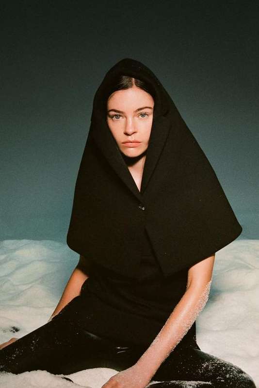 Fashion portrait of a woman wearing a black hooded garment, seated in a snowy setting against a muted blue background.