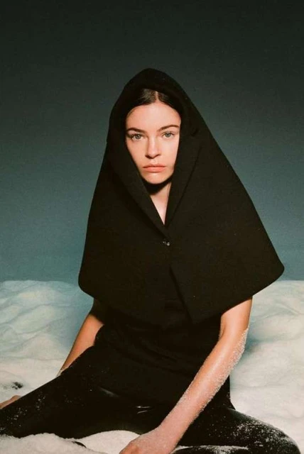 Fashion portrait of a woman wearing a black hooded garment, seated in a snowy setting against a muted blue background.