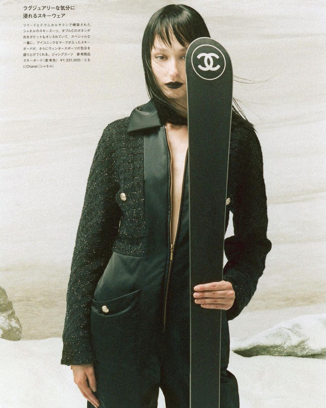 Fashion shot of a slender adult female in a black jumpsuit and jacket, holding a black Chanel ski, posed against a neutral backdrop.