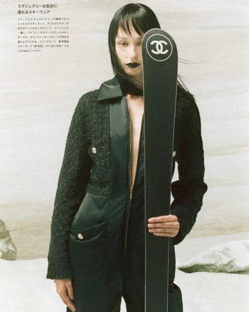 Fashion shot of a slender adult female in a black jumpsuit and jacket, holding a black Chanel ski, posed against a neutral backdrop.