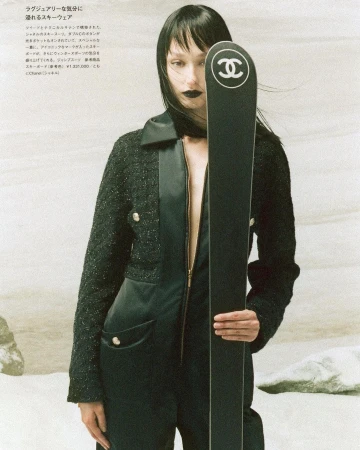 Fashion shot of a slender adult female in a black jumpsuit and jacket, holding a black Chanel ski, posed against a neutral backdrop.