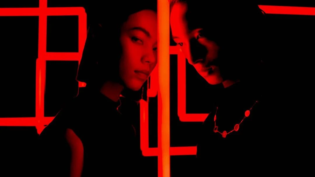 Dramatic studio portrait of a stylish young adult woman and man, with red grid lighting in a high-contrast, dark fashion aesthetic.
