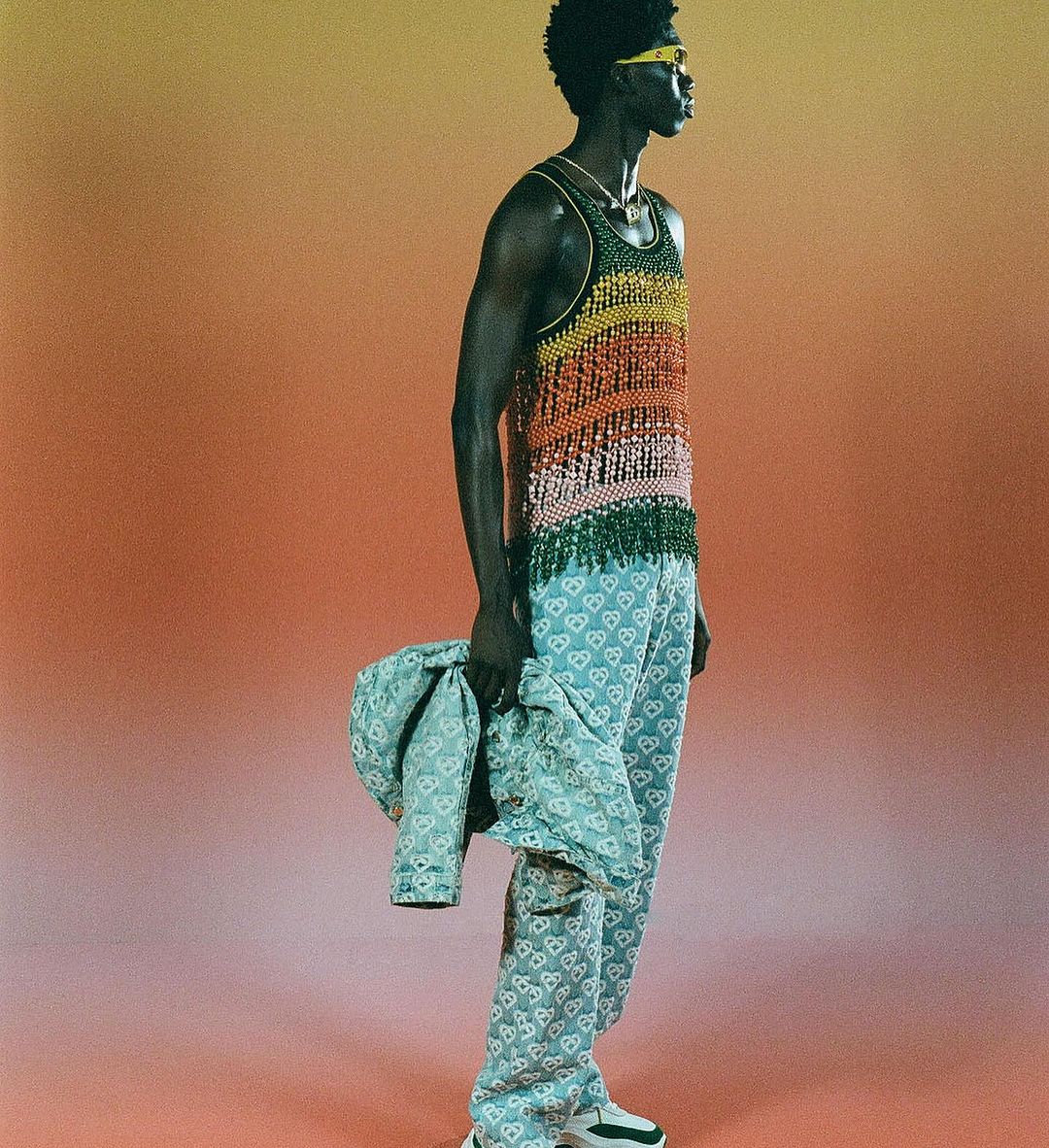 Dark-skinned male fashion model in patterned pants and colorful fringed top poses in a studio against an orange gradient background.