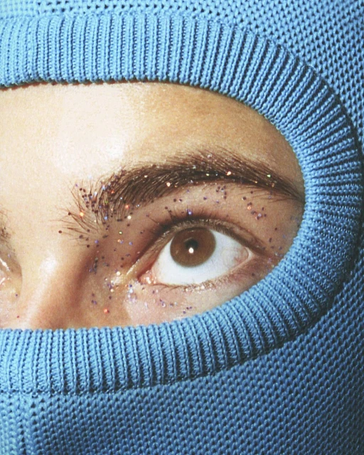 Close-up shot of a person's eye and face partially covered by a light blue knitted balaclava, with glitter on their brow and under their eye.