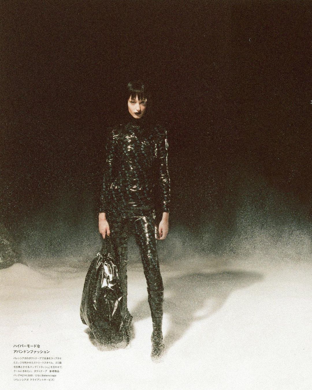 A woman poses in a shiny black outfit with a black trash bag in a dark environment, creating a striking fashion editorial image.