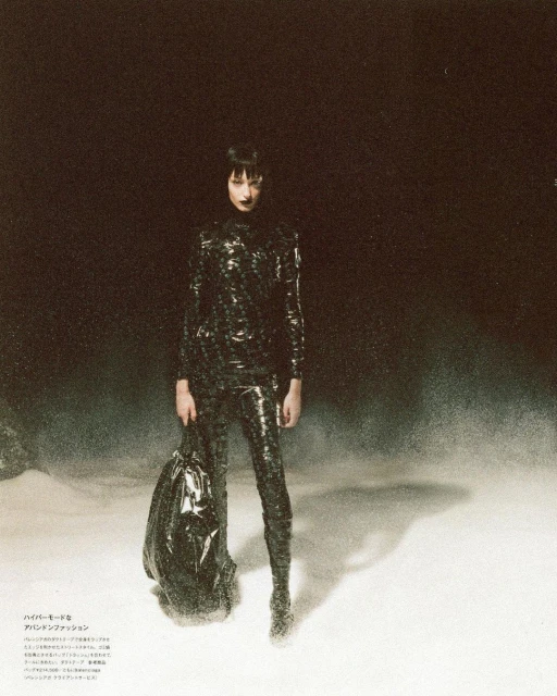 A woman poses in a shiny black outfit with a black trash bag in a dark environment, creating a striking fashion editorial image.