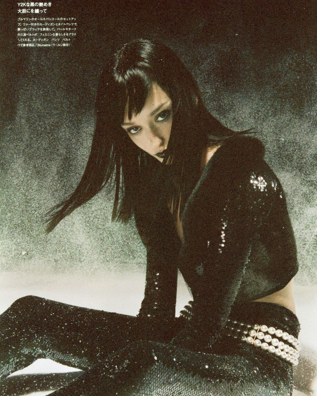 Elegant woman with dark hair poses indoors wearing black sequined pants and a matching long-sleeved top, accessorized with a pearl belt.