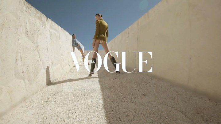 Two fashion models strike poses in a concrete environment under a clear sky. One model stands facing forward, while the other leans against the wall.