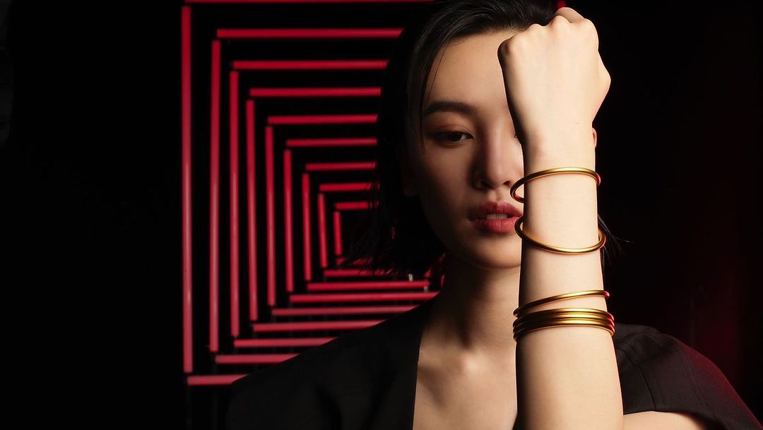 Close-up of an Asian woman wearing gold bracelets in front of a geometric red light pattern on a dark background in a studio shot.