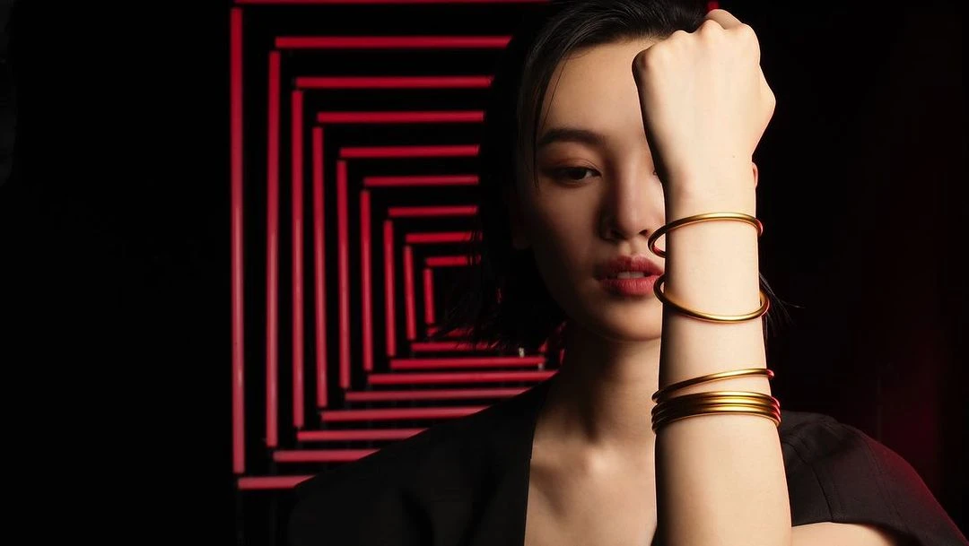 Close-up of an Asian woman wearing gold bracelets in front of a geometric red light pattern on a dark background in a studio shot.