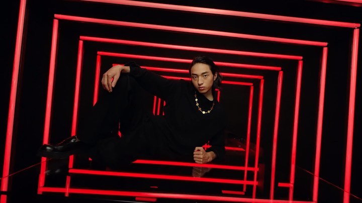 Young man in black clothing poses against a backdrop of neon red lights that create a square tunnel effect in a studio environment.