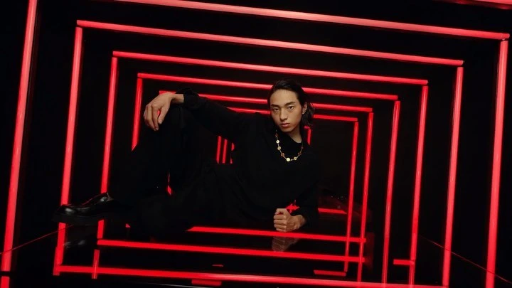 Young man in black clothing poses against a backdrop of neon red lights that create a square tunnel effect in a studio environment.