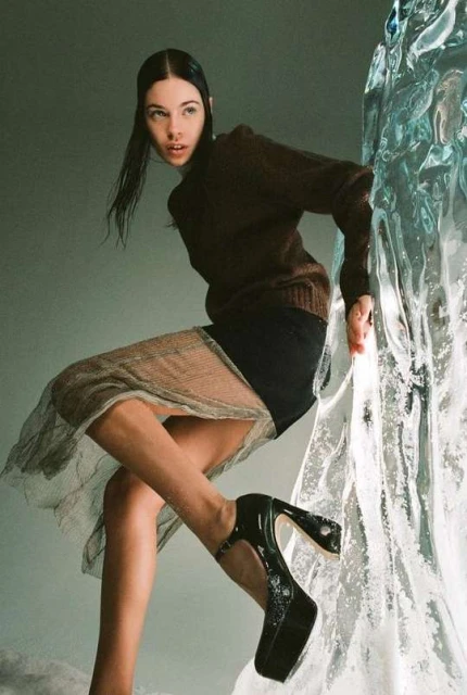 Woman poses in a brown sweater, black skirt and mesh overlay, and black platform heels next to a textured glass structure.