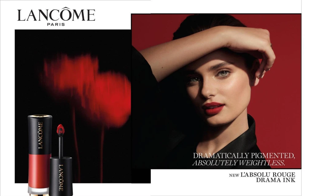 Lancome beauty shot featuring an adult woman with red lipstick and red lip product on a black background, product on the left.