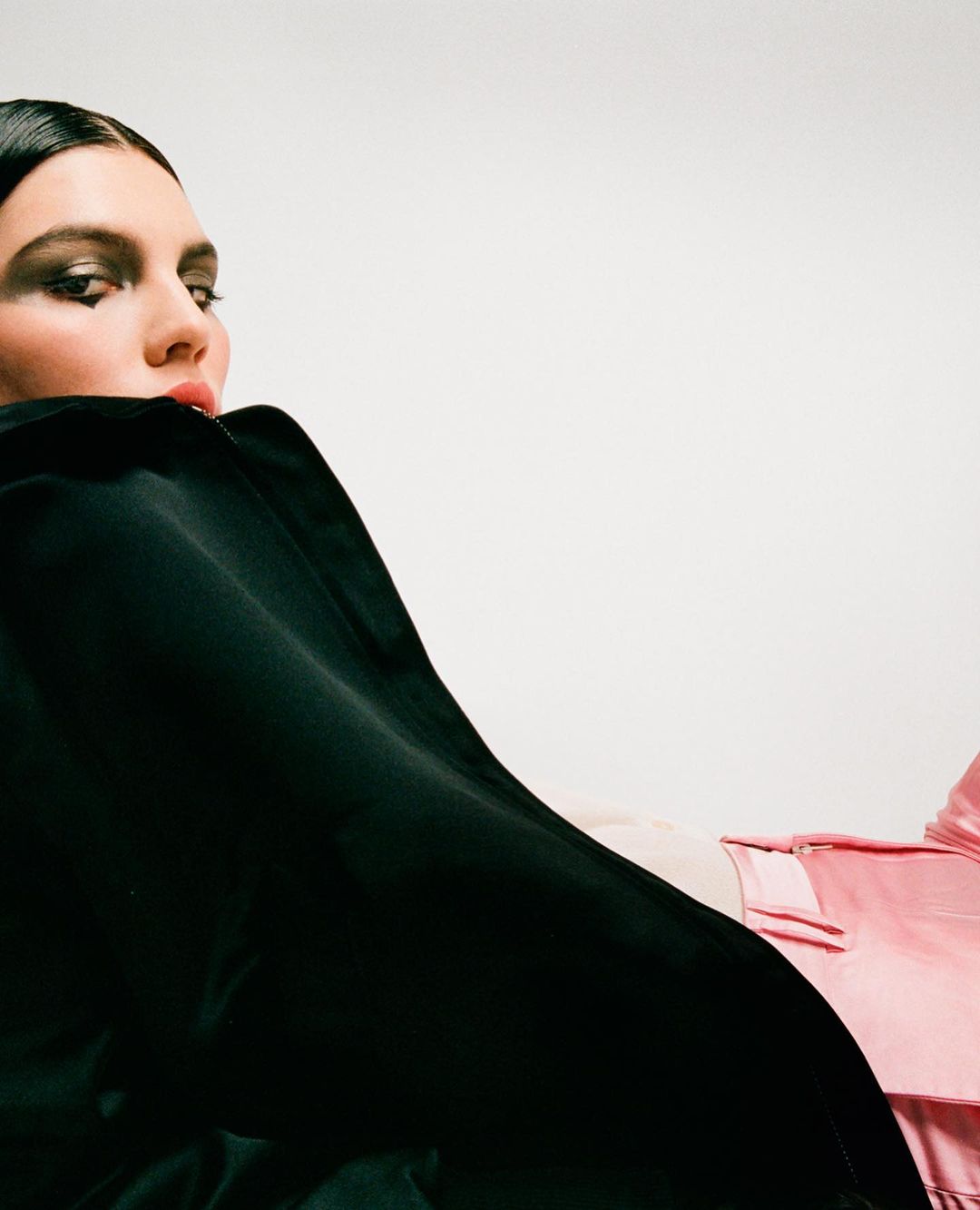 Portrait of a woman with dark hair and dramatic eye makeup wearing a black top and pink satin pants against a white backdrop.