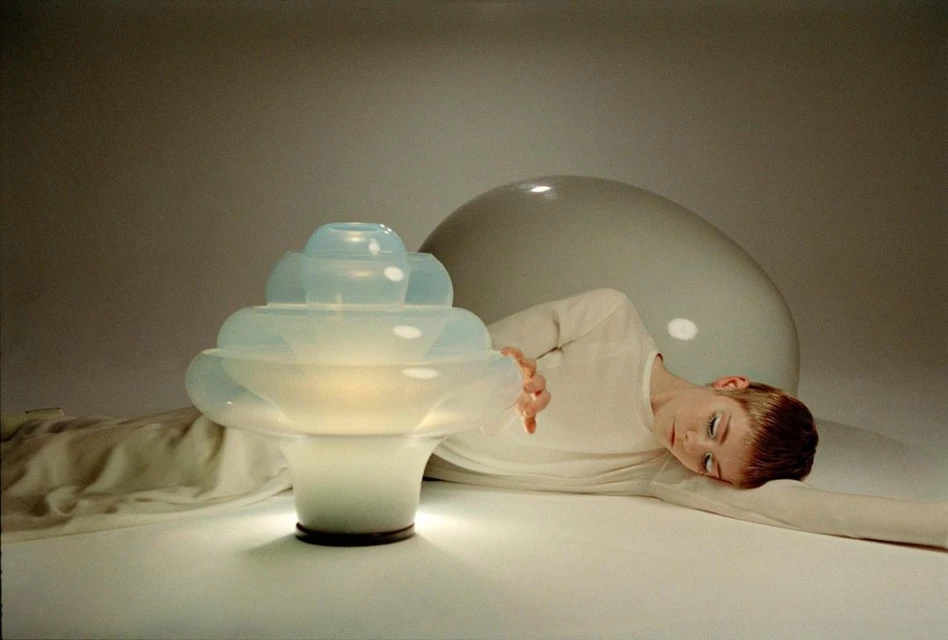 Woman in white next to an illuminated sculptural table lamp and large white sphere in a softly lit interior, modern and minimalist.